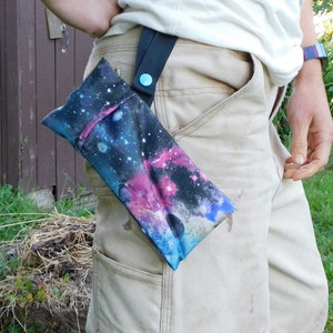 May include: A fabric phone case with a galaxy print in shades of blue, purple, and pink. The case is attached to a black strap with a blue button closure. The strap is connected to a belt loop.