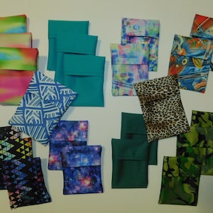 May include: A collection of 14 fabric pouches in various patterns and colors, including green, blue, pink, black, white, and camouflage. The pouches are made of different fabrics, including cotton, linen, and silk. They are all folded and stacked on a white surface.