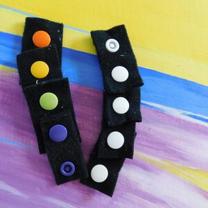 May include: A set of black fabric strips with colorful and white buttons. The buttons are attached to the fabric strips with a loop and hook fastener.
