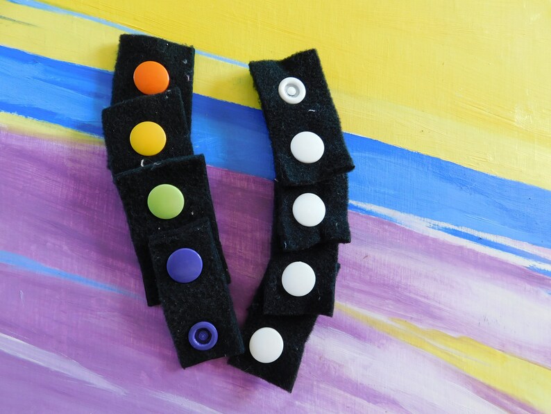 May include: A set of black fabric strips with colorful and white buttons. The buttons are attached to the fabric strips with a loop and hook fastener.