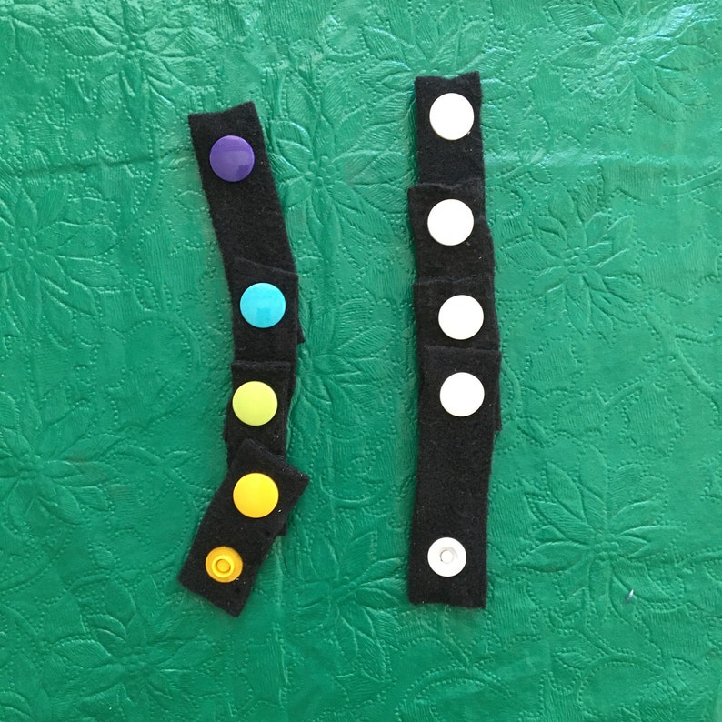 May include: Two black fabric strips with rows of colorful buttons. The strip on the left has purple, blue, green, yellow, and yellow buttons. The strip on the right has white buttons.