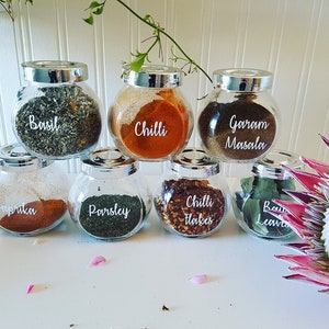 May include: Seven glass spice jars with silver lids and white labels. The labels are handwritten with the names of different spices: Basil, Chilli, Garam Masala, Paprika, Parsley, Chilli Flakes, and Bay Leaves.
