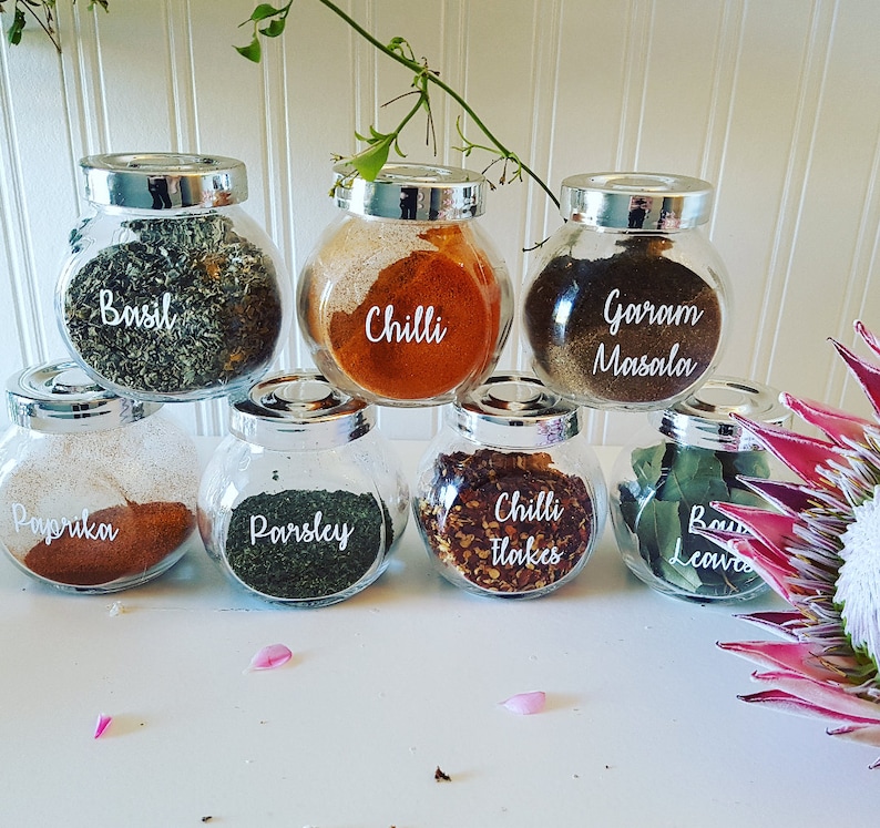 K&ouml;nnte beinhalten: Sieben gl&auml;serne Gew&uuml;rzgl&auml;ser mit silbernen Deckeln und wei&szlig;en Etiketten. Die Etiketten sind handgeschrieben mit den Namen verschiedener Gew&uuml;rze: Basilikum, Chili, Garam Masala, Paprika, Petersilie, Chiliflocken und Lorbeerbl&auml;tter.