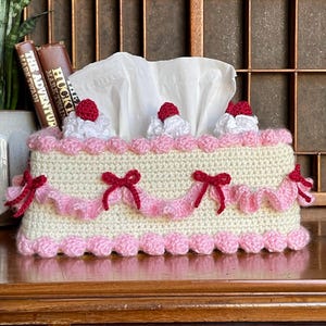May include: A cream-coloured crocheted tissue box cover, designed to look like a cake. It has pink trim, bows, and decorative elements. The top features white frosting and red cherry accents. The tissue box is on a wooden surface.