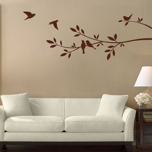 May include: A brown silhouette of a tree branch with birds perched on the branches. The tree branch is on a light brown wall with a white sofa in the foreground.
