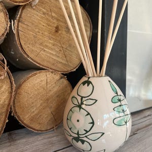 Può includere: Un diffusore di oli essenziali in ceramica bianca con un design floreale verde e turchese, riempito di bastoncini di legno. Il diffusore è posizionato su una superficie di legno, con tronchi impilati sullo sfondo.