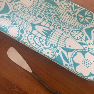 May include: A white ceramic serving tray with a teal floral pattern and a silver butter knife with a black twisted handle.