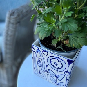 Puede incluir: Una maceta cuadrada de cerámica blanca con diseños azules pintados a mano, que incluyen peces y motivos florales, contiene una planta verde frondosa. La maceta está sobre una superficie blanca, con un fondo borroso.