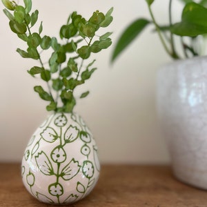 Op de afbeelding: Een witte keramische vaas met een groen bloemenpatroon, met een takje groene, bladerrijke planten. De vaas heeft de vorm van een traan en staat op een houten oppervlak.