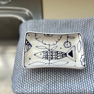 May include: A small white ceramic dish with a blue and white floral design and a fish in the centre. The dish is sitting on a blue and white patterned fabric.