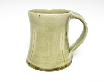 May include: A handmade, light green ceramic mug with a curved body and a sturdy handle. The mug has a slightly textured surface and a dark brown base. The design is simple and elegant, suitable for everyday use.
