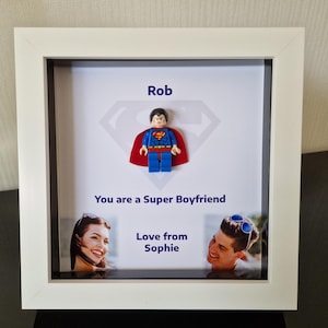 Op de afbeelding: Een wit fotolijstje met een Superman Lego minifiguur erin. Het lijstje heeft een witte passe-partout en een glazen voorzijde. De tekst "Rob" staat bovenaan het lijstje, en de tekst "You are a Super Boyfriend" staat onder de minifiguur. De tekst "Love from Sophie" staat onderaan het lijstje.