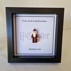 Może przedstawiać: Czarny obraz w ramie z białym tłem. Obraz przedstawia minifigurkę Lego Dumbledore'a z serii Harry Potter. Tekst "Nie warto rozpamiętywać snów... I zapominać o życiu" jest nadrukowany na obrazie.