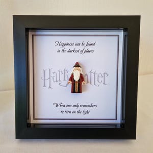 Può includere: Un'opera d'arte incorniciata con una figura in miniatura di Silente di Harry Potter. Il testo recita: "La felicità può essere trovata nei luoghi più oscuri" e "Quando ci si ricorda solo di accendere la luce". La cornice è nera.