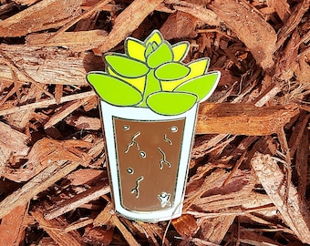 May include: A gold-tone enamel pin featuring a green succulent plant in a brown pot. The pin is on a background of brown wood chips.