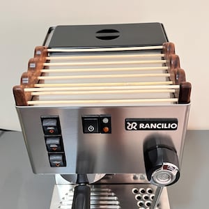 Op de afbeelding: Een roestvrijstalen Rancilio espressomachine met een kopjeswarmer voorzien van een houten rek. Het voorpaneel bevat drie zwarte tuimelschakelaars, een aan-uitknop met indicatielampje en een zwarte stoomknop met een zilveren midden.