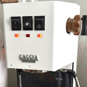 May include: A white Gaggia Classic espresso machine with wooden detailing. It has three black switches, indicator lights, and the Gaggia Classic logo. The machine includes a steam wand and a portafilter for making coffee.