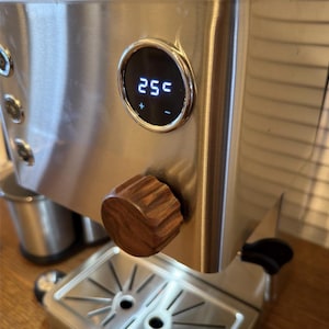 May include: Stainless steel espresso machine with a wooden knob and digital display showing "255". The machine has a silver finish and a detailed design. The display has plus and minus buttons.