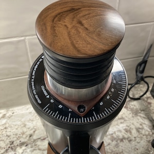 May include: A close-up of a black and silver coffee grinder with a wooden top. The grinder has a dial that goes from 0 to 90, with markings for 75, 85, and 90. The dial is labeled "COARSE" and has a warning that says "Do not adjust past "0" to prevent burr damage."