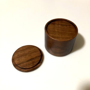 Peut inclure: Une boîte ronde en bois avec un couvercle. La boîte est en bois brun foncé et a une finition lisse. Le couvercle est légèrement surélevé par rapport à la boîte et a un bord légèrement arrondi.