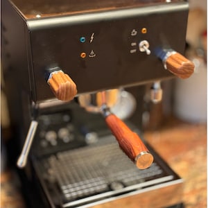 Peut inclure: Une machine à espresso noire avec un dessus en bois et des poignées en bois. La machine possède un écran numérique et plusieurs boutons. La machine est posée sur un comptoir.