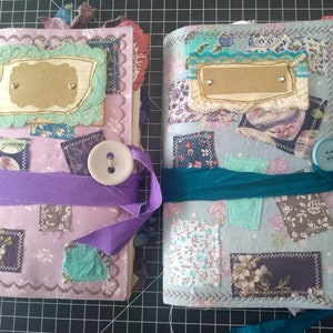 Op de afbeelding: Twee handgemaakte dagboeken met stoffen omslagen. Het dagboek links heeft een paarse en roze omslag met een groene en witte kantversiering. Het dagboek rechts heeft een blauwe en witte omslag met een blauwe en witte kantversiering.