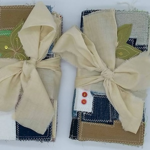 May include: Two handmade journals wrapped in fabric with a white ribbon bow. The fabric is a patchwork of denim, brown canvas, and white cotton. Each journal has a green fabric flower with gold sequins.