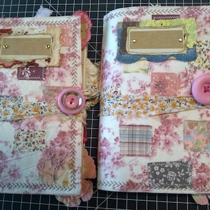 May include: Two handmade journals with pink floral fabric covers, each with a brown paper label and pink buttons. The journals are decorated with lace, ribbon, and fabric scraps.