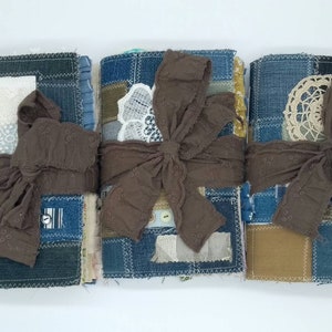 Puede incluir: Tres diarios hechos a mano con tapas de mezclilla y tela, atados con una cinta marrón. Cada diario tiene un diseño diferente con encaje, recortes de tela y costuras.