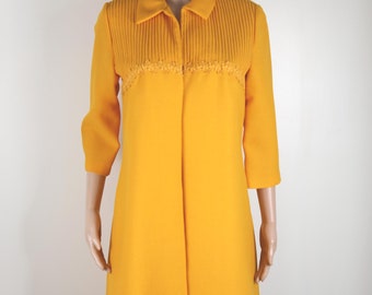 Pode incluir: Um vestido amarelo com fecho de botões na frente e uma guarnição de renda em volta do decote. O vestido tem um corpete plissado e uma saia ligeiramente rodada.