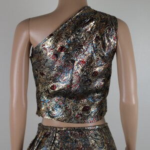 May include: A two-piece outfit with a gold, red, and blue patterned design. The top is a one-shoulder crop top and the skirt is a high-waisted skirt.