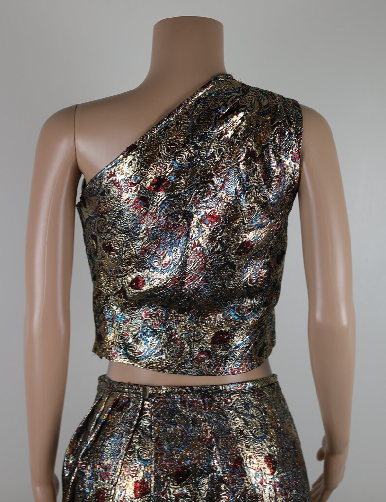 May include: A two-piece outfit with a gold, red, and blue patterned design. The top is a one-shoulder crop top and the skirt is a high-waisted skirt.