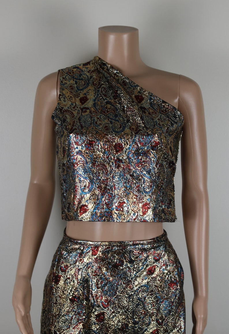May include: A two-piece outfit with a gold, blue, and red paisley pattern. The top is a one-shoulder crop top and the bottom is a skirt.