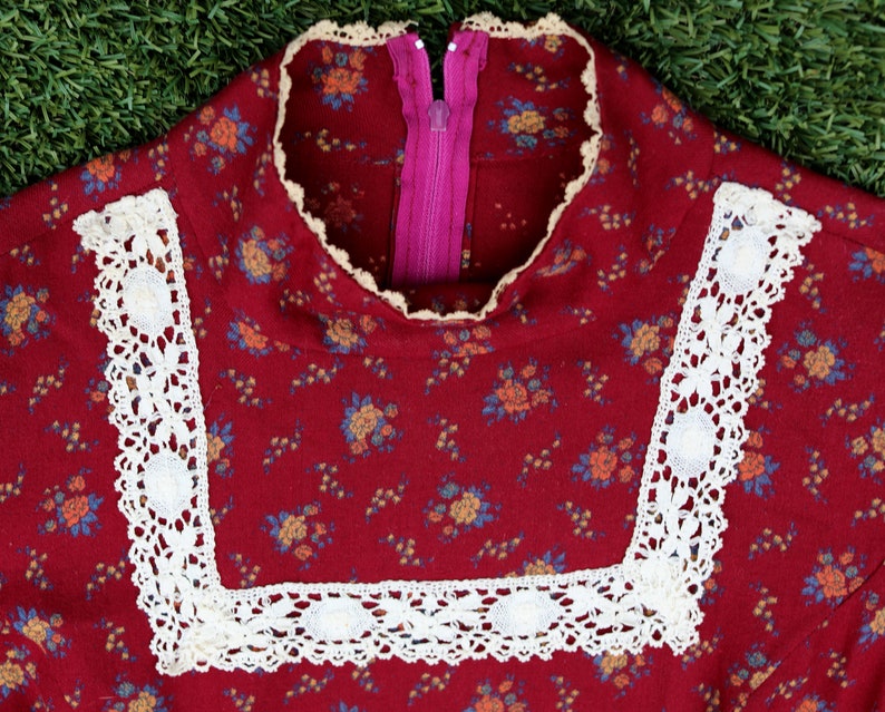 May include: Close-up of a red floral dress with white lace trim around the neckline. The dress has a pink zipper in the back.