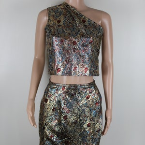 May include: A two-piece outfit with a gold, blue, and red brocade pattern. The top is a one-shoulder crop top and the skirt is a pencil skirt.
