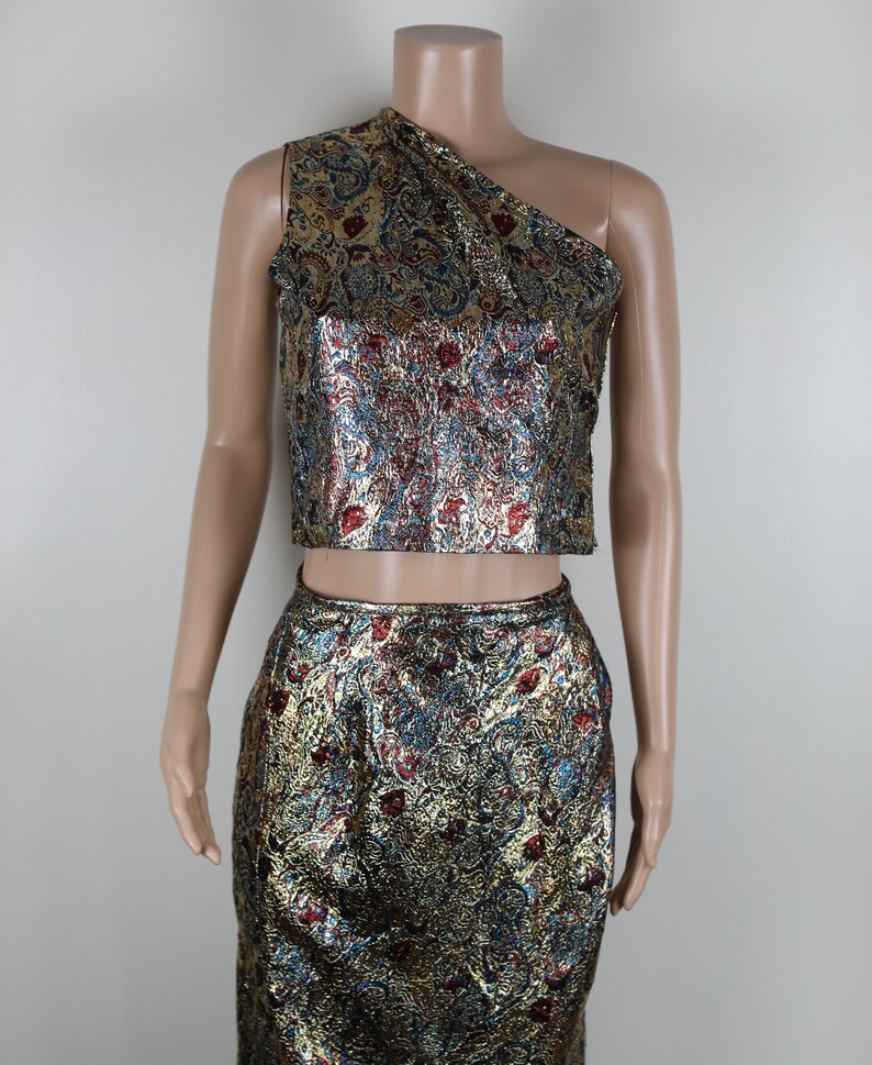 May include: A two-piece outfit with a gold, blue, and red brocade pattern. The top is a one-shoulder crop top and the skirt is a pencil skirt.