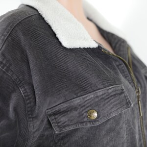 May include: A dark gray corduroy jacket with a white fleece collar and a zipper closure. The jacket has a large pocket on the front with a metal button.