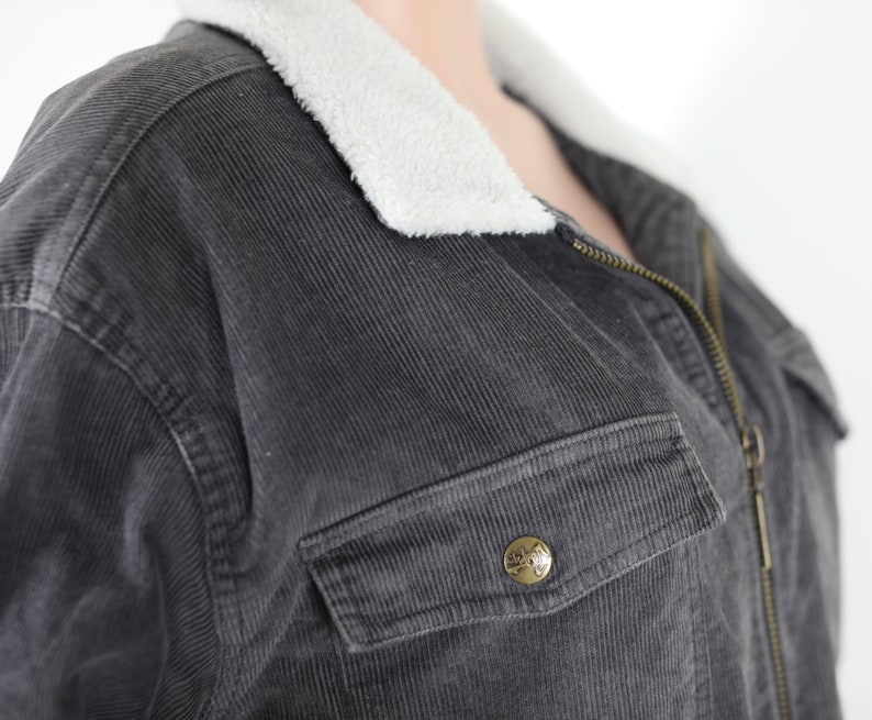 May include: A dark gray corduroy jacket with a white fleece collar and a zipper closure. The jacket has a large pocket on the front with a metal button.