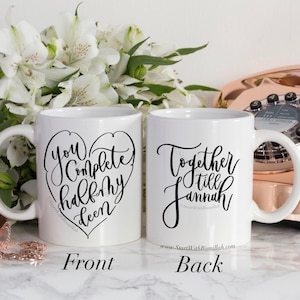May include: Two white ceramic mugs with black calligraphy. The front mug says "You complete half my deen" in a heart shape. The back mug says "Together till Jannah" with a website address below.
