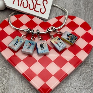 May include: A silver charm bracelet with five book-shaped charms. Each charm features a book cover design with the title and author's name. The charms are suspended from the bracelet and are displayed on a red and white checkered heart-shaped tray.