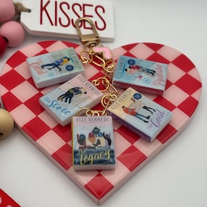 May include: A gold keychain with miniature book charms. Each charm features a book cover from the "Elle Kennedy" series, including "The Deal," "The Score," "The Legacy," "The Goal," and "The Mistake." The keychain is attached to a pink heart-shaped base with a red and white checkered pattern.