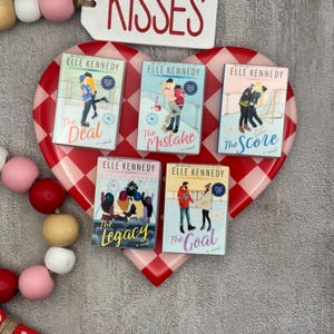 May include: Five books by Elle Kennedy are displayed on a red and white heart-shaped tray. The book titles are "The Deal", "The Mistake", "The Score", "The Legacy", and "The Goal". The word "Kisses" is written on a white tag above the books.