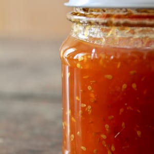 May include: A glass jar filled with a bright orange, seed-filled jam. The jar has a white lid and is sitting on a wooden surface.