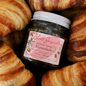 May include: A glass jar of Sweet Jaquelina Heirloom Tomato Marmalade with a pink label. The jar is surrounded by golden brown croissants.