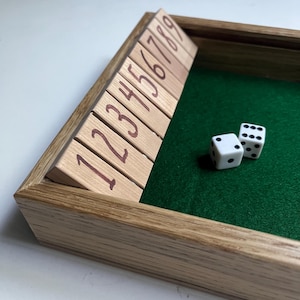 May include: Wooden dice game set with a green felt interior. The set includes numbered wooden tiles and two white dice with black dots. The wooden frame has a natural finish.