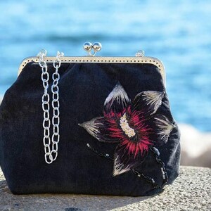 May include: A black velvet clutch bag with a silver chain strap and an embroidered flower design. The flower is pink, white, and black.