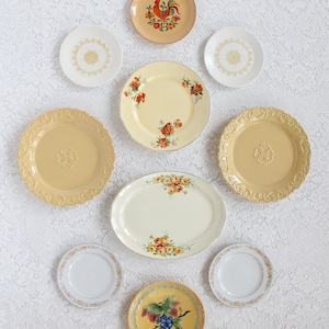 Może przedstawiać: Kolekcja dziewięciu vintage ceramicznych talerzy z różnymi wzorami kwiatowymi i owocowymi. Talerze są w odcieniach żółtego, białego i pomarańczowego. Niektóre talerze mają podniesioną krawędź i ozdobną ramkę.