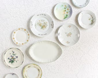May include: A collection of vintage decorative plates with floral and botanical designs. The plates vary in size and shape, including round and oval. Some plates feature text, and the color palette includes white, yellow, green, and purple.
