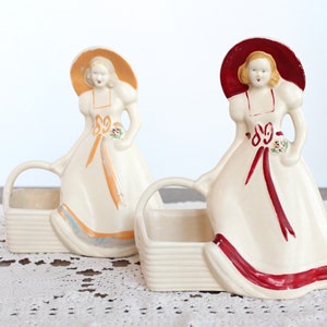 May include: Two vintage ceramic planter figurines of women in dresses and hats holding baskets. One woman is wearing a red hat and dress with a white trim, the other is wearing a yellow hat and dress with a blue trim.
