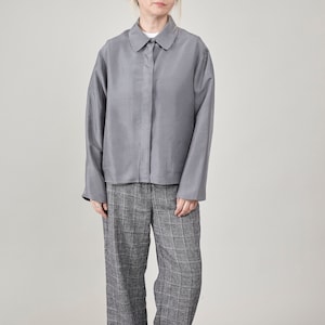 May include: A person wearing a grey collared shirt and grey plaid trousers. The shirt has a button-down front and two front pockets. The trousers are wide-legged and feature a grey and white plaid pattern. The outfit is paired with brown shoes.